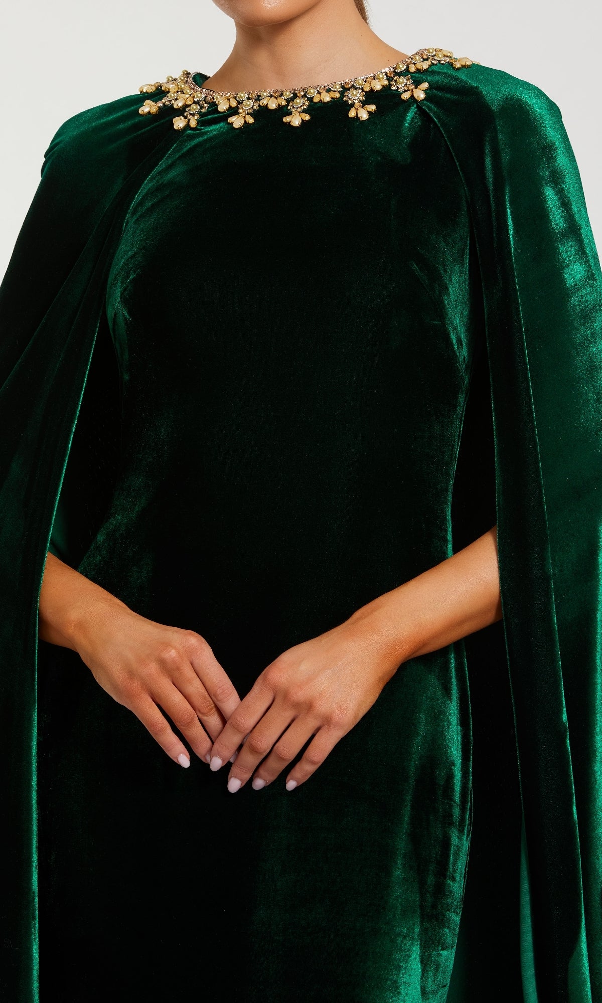 A person wearing the Mac Duggal 68977 Green Velvet Caped Formal Dress with gold embellishments around the neckline, hands clasped in front.